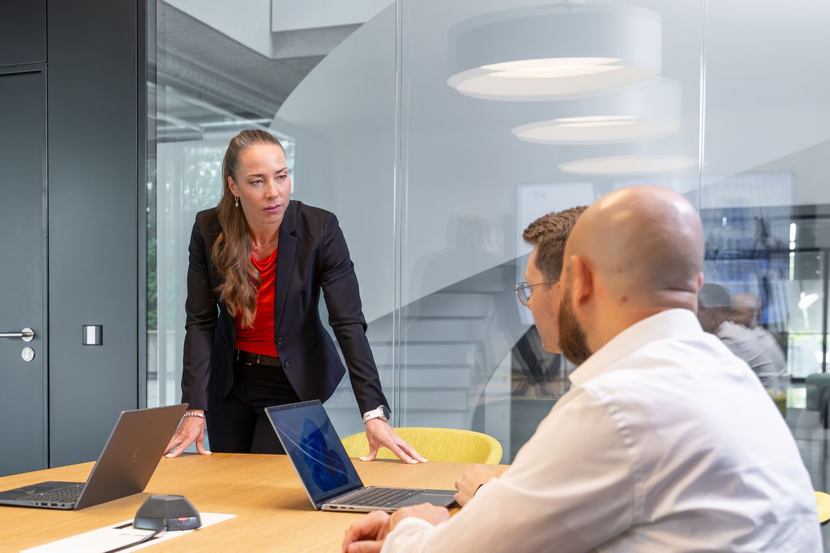 Beate van Loo-Born standing at a boardroom table, speaking to two colleagues seated opposite her.